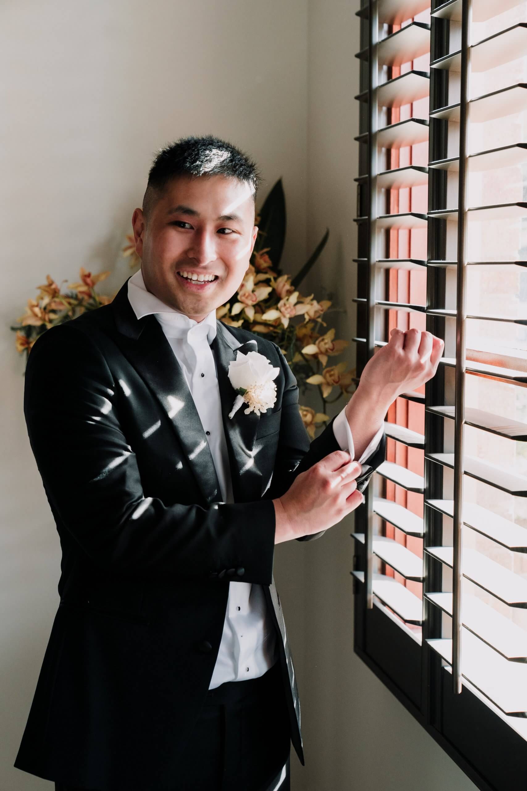 Groom standing by a window and adjusting his cuffs with a smile